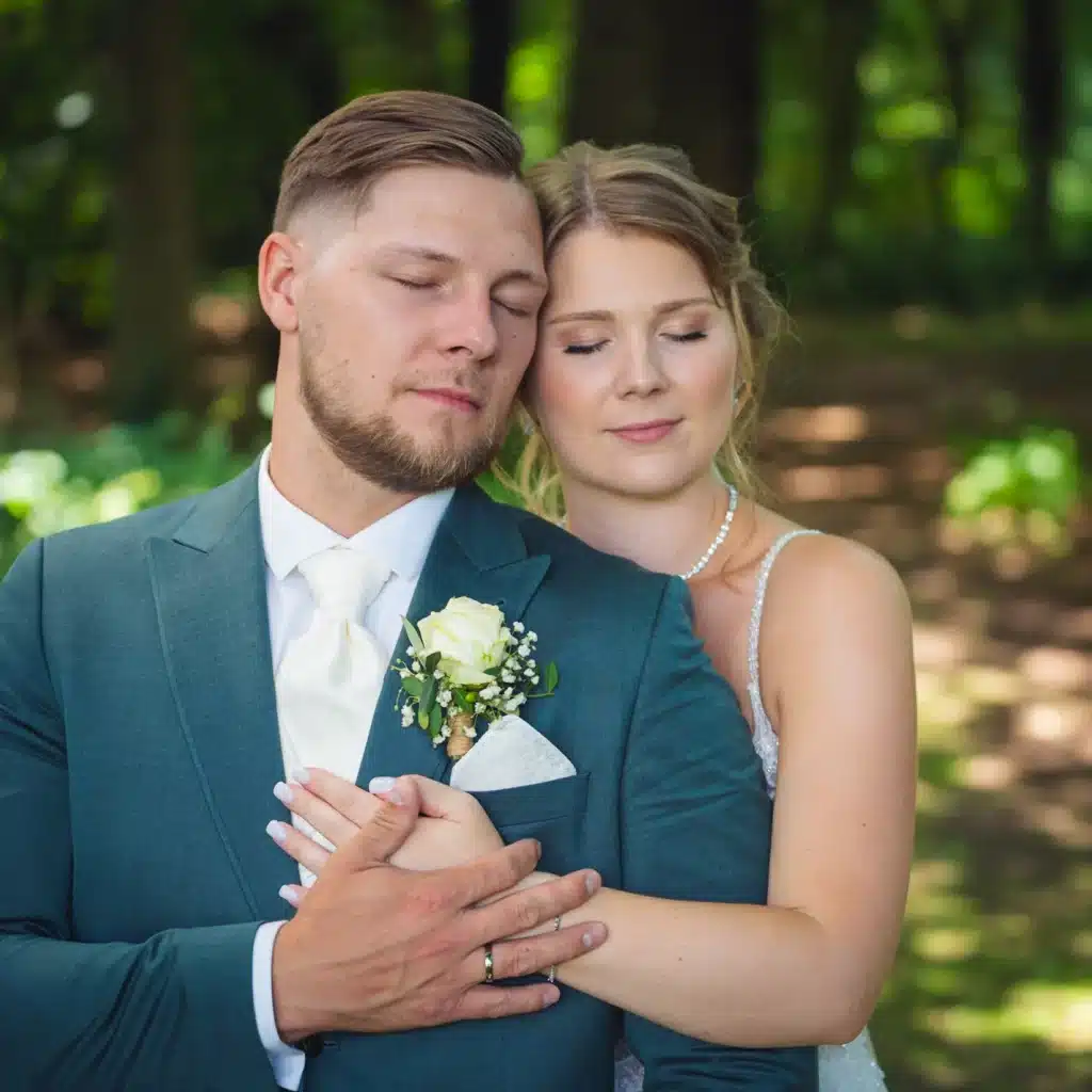 Ein Bräutigam in einem grünen Anzug und eine Braut in einem weißen Kleid umarmen sich mit geschlossenen Augen und strahlen Ruhe und Glück aus.