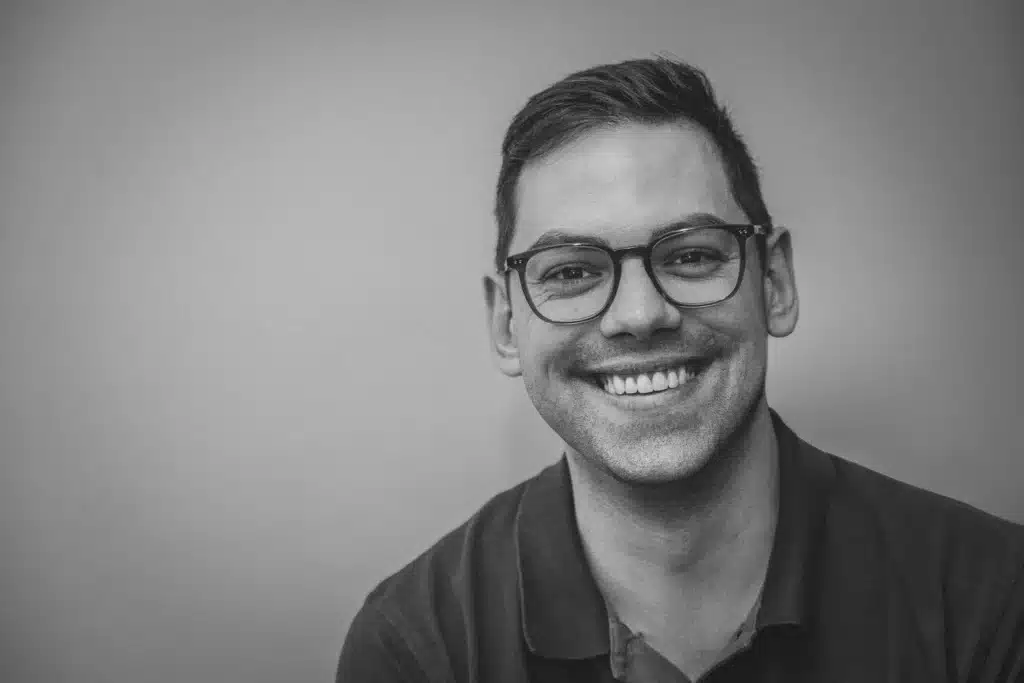 Sebastian als lächelnder Mann mit Brille, kurzem Haar und einem Poloshirt, aufgenommen in Schwarz-Weiß.