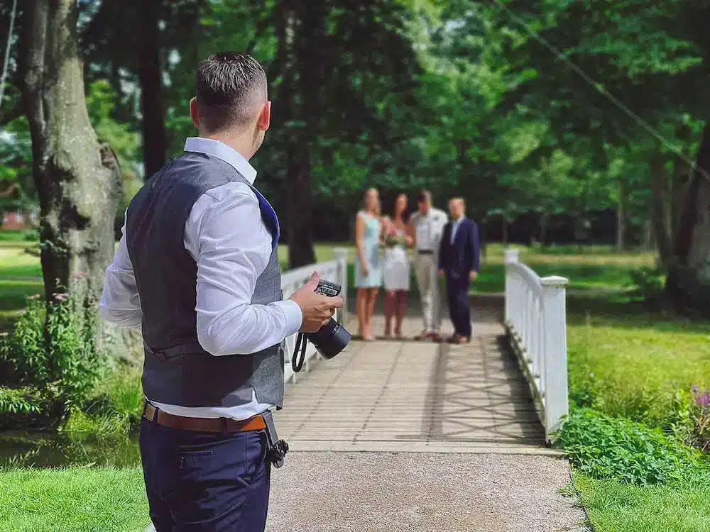 Sebastian Selle in einem grauen Anzug fotografiert eine Hochzeitsgesellschaft, die auf einer weißen Brücke in einem grünen Park am Schloss Evenburg posiert.